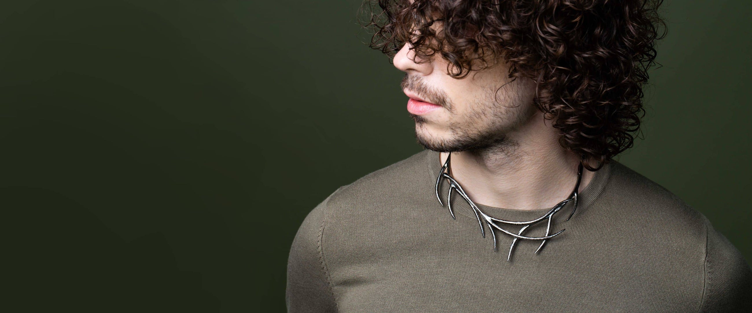 Homme dont le visage est caché par ses cheveux et portant un collier en forme de branches sur un pull kaki.